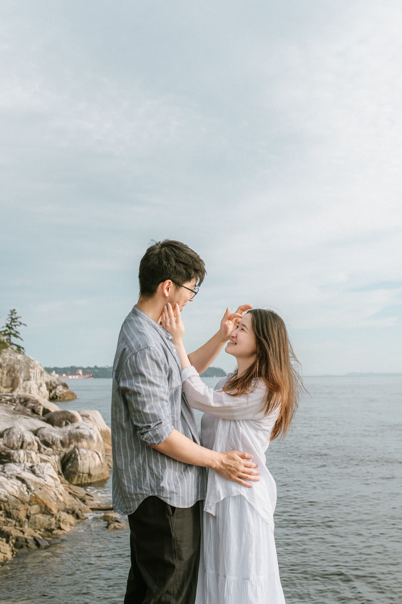 Couple beach photography, a couple embrace on a rocky shore by the sea, she in a white dress with her hand on his cheek, soft overcast light