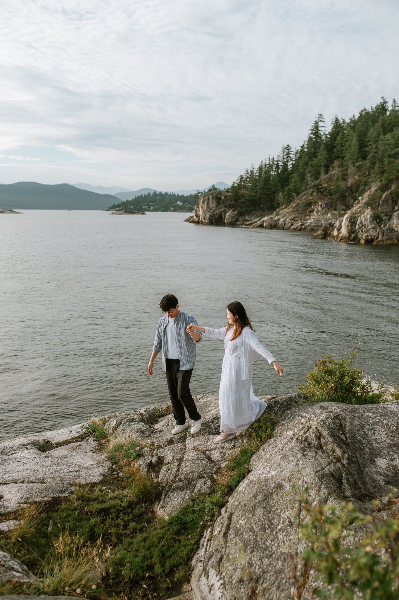 Woods engagement photos, a couple walk hand in hand over the rocks along a forested shoreline, islands in the distance