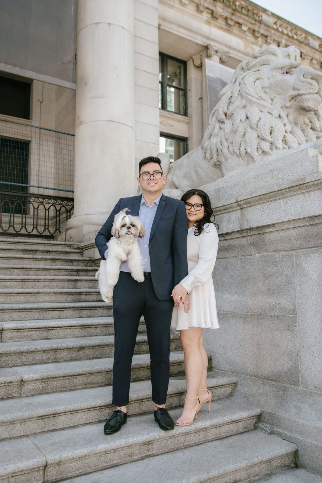 Creative couple photography, a couple with a small white dog in his arms stand on the stone steps of a grand neoclassical building beside a carved stone lion