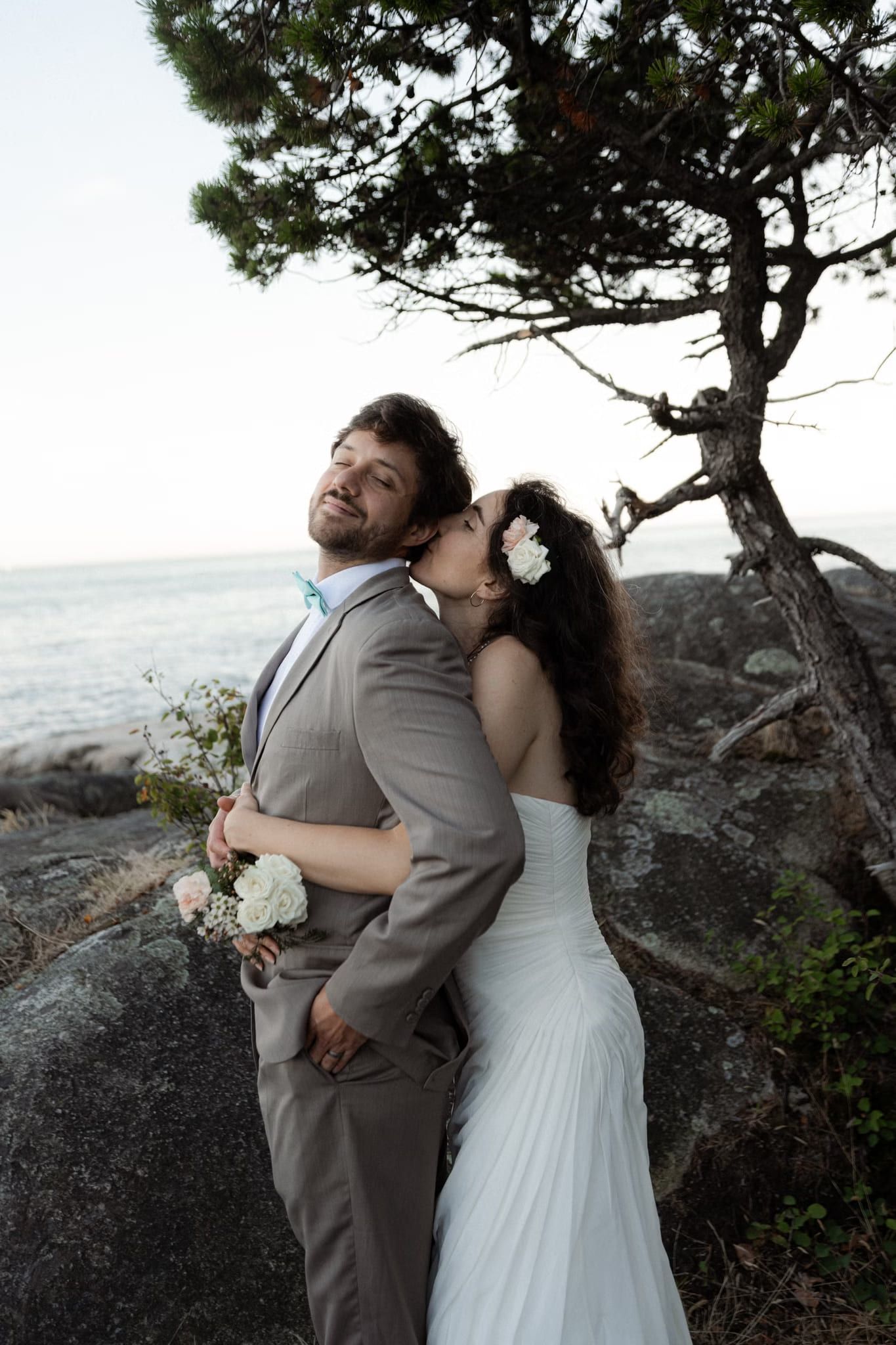 Bride kissing the groom's cheek beneath a windswept pine on the rocky shore