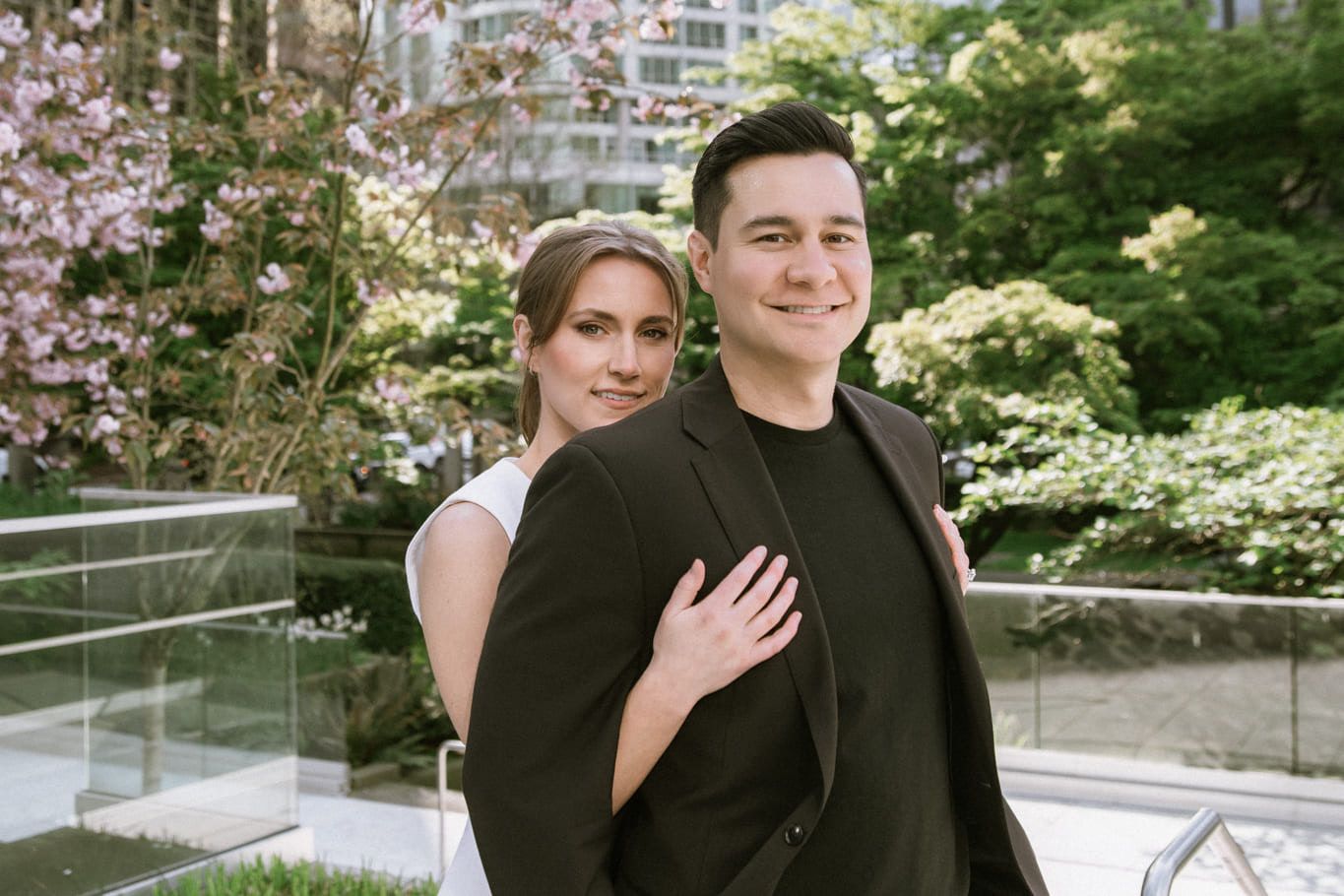 Candid engagement photos, a couple in a black blazer and a white top smile among cherry blossoms on a rooftop terrace, city buildings behind