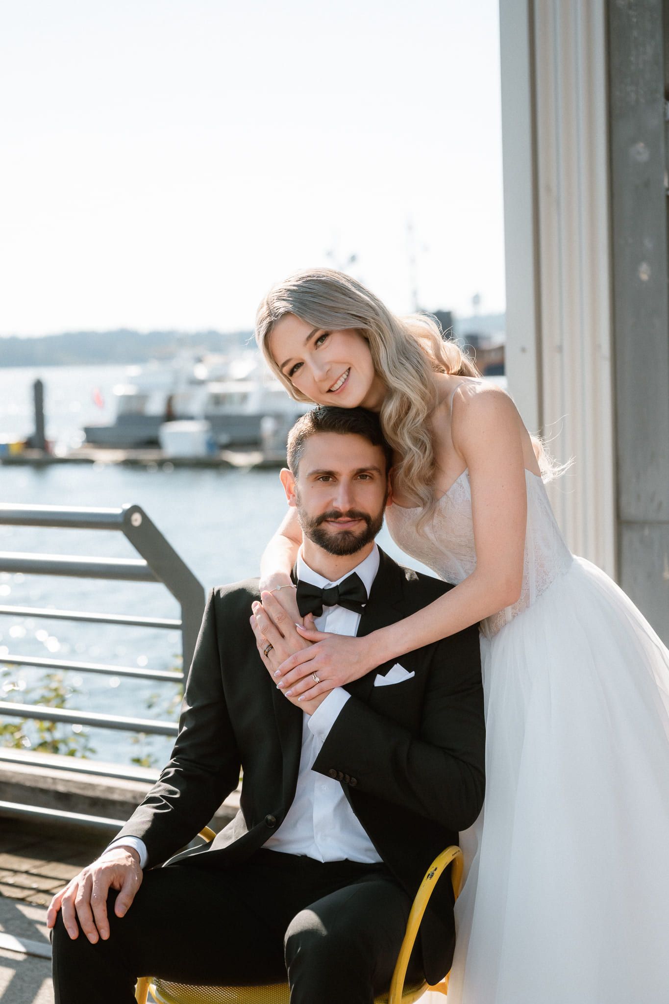 Groom photos on a waterfront terrace, bride embracing the groom from behind