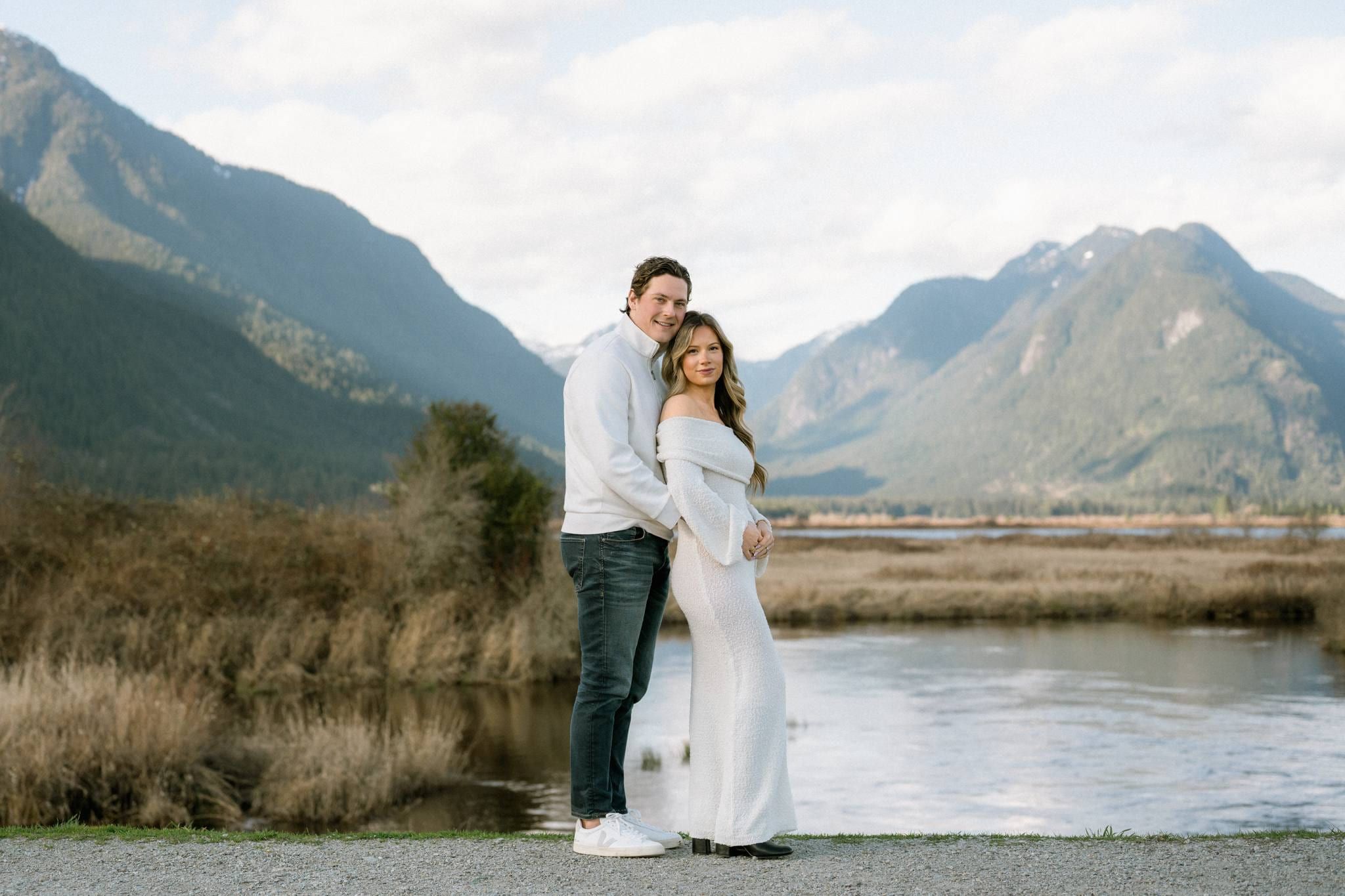 Expectant couple in white by the water with snow-capped mountains behind