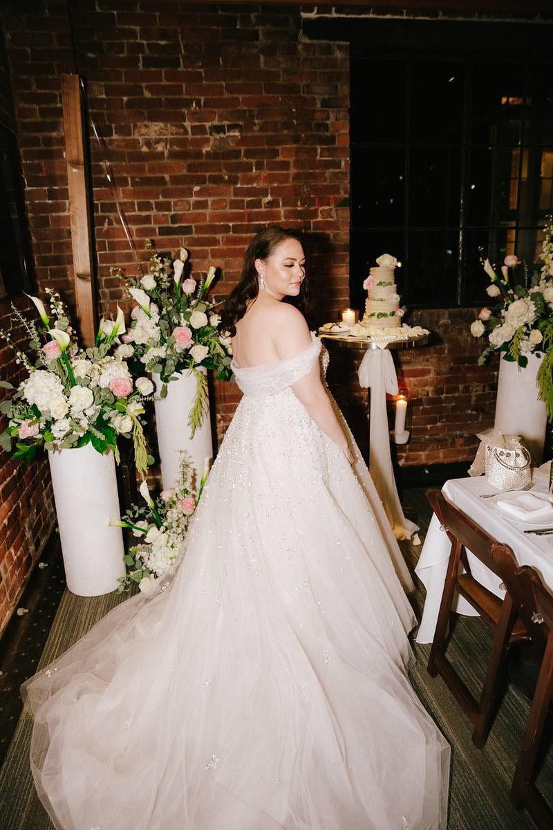 Wedding gown photos, bride in a full tulle gown admiring the cake in a brick-walled venue