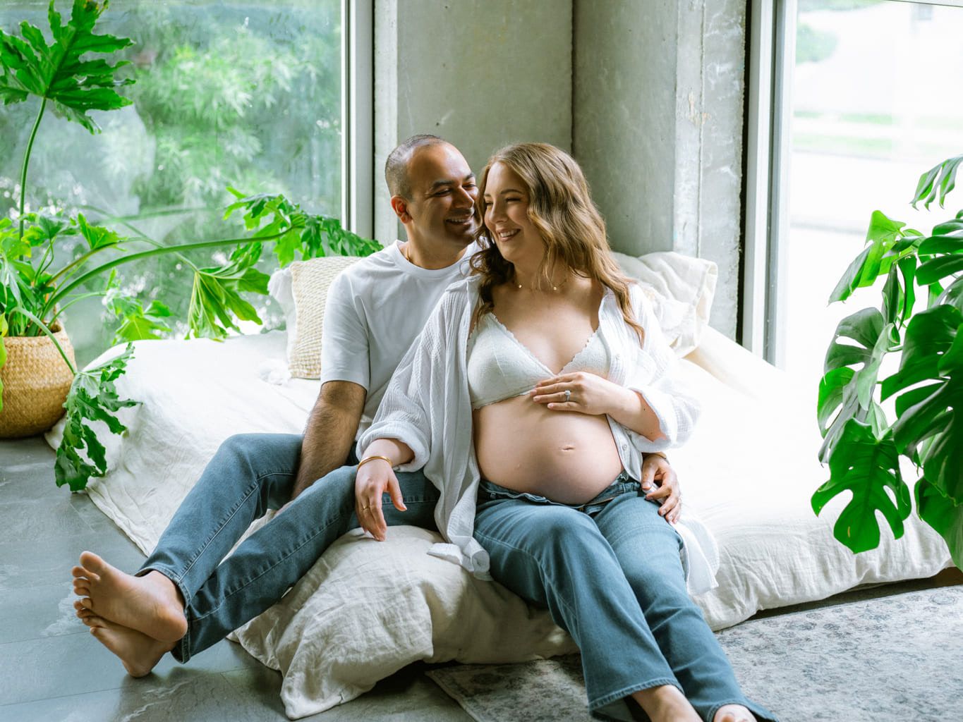 Indoor studio maternity photoshoot, expectant couple relaxing on cushions by a big studio window with plants