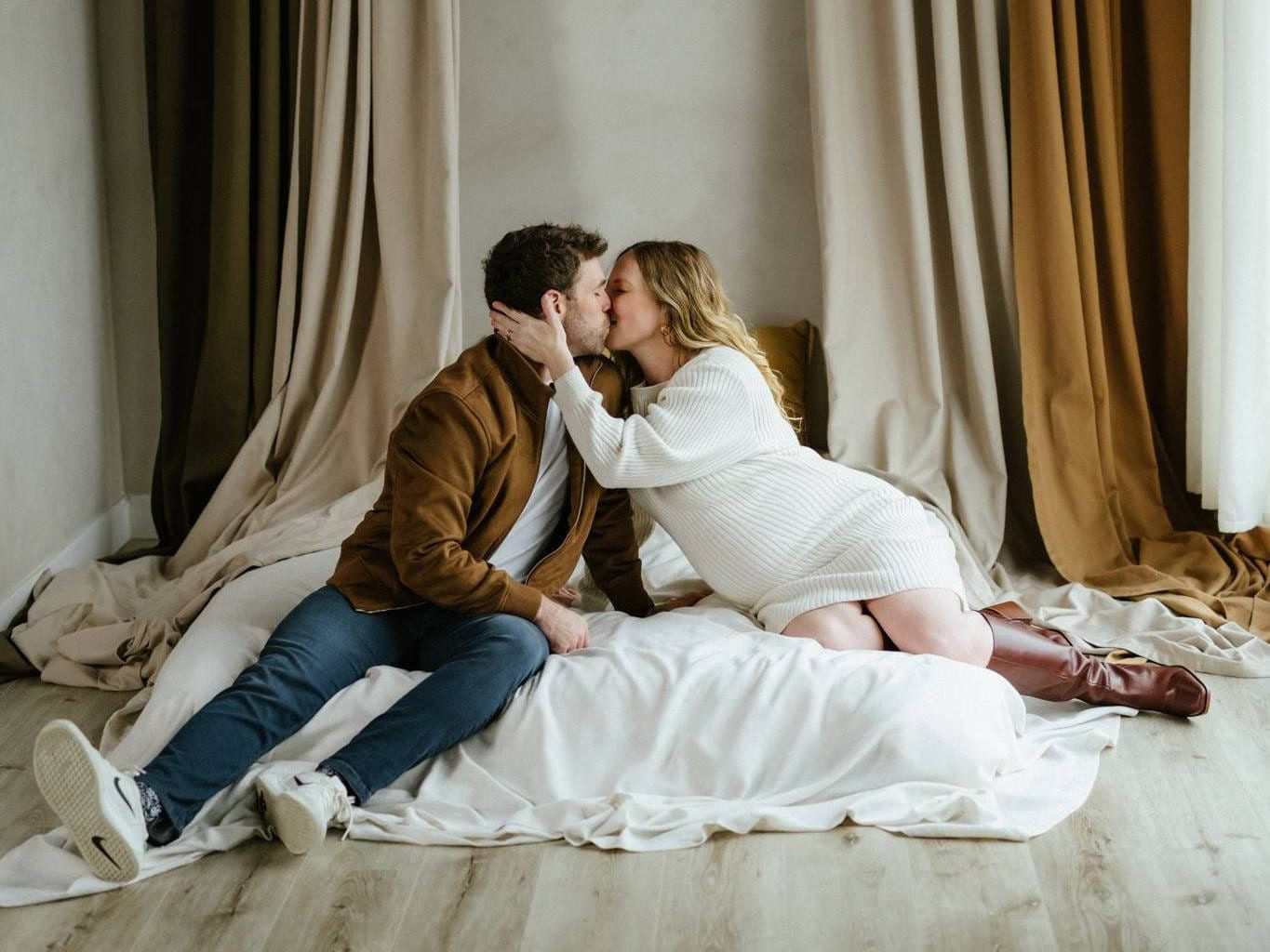 Photography studio session, couple kissing on a white bed between draped curtains in a studio