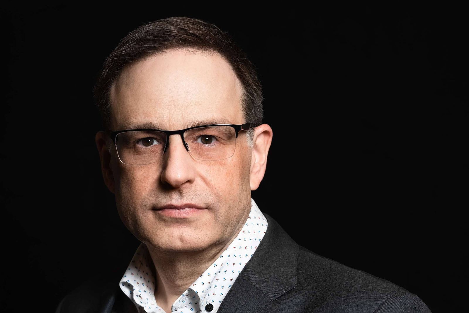 Headshot photo studio session, professional headshot of a man in glasses and a dark blazer on a black backdrop