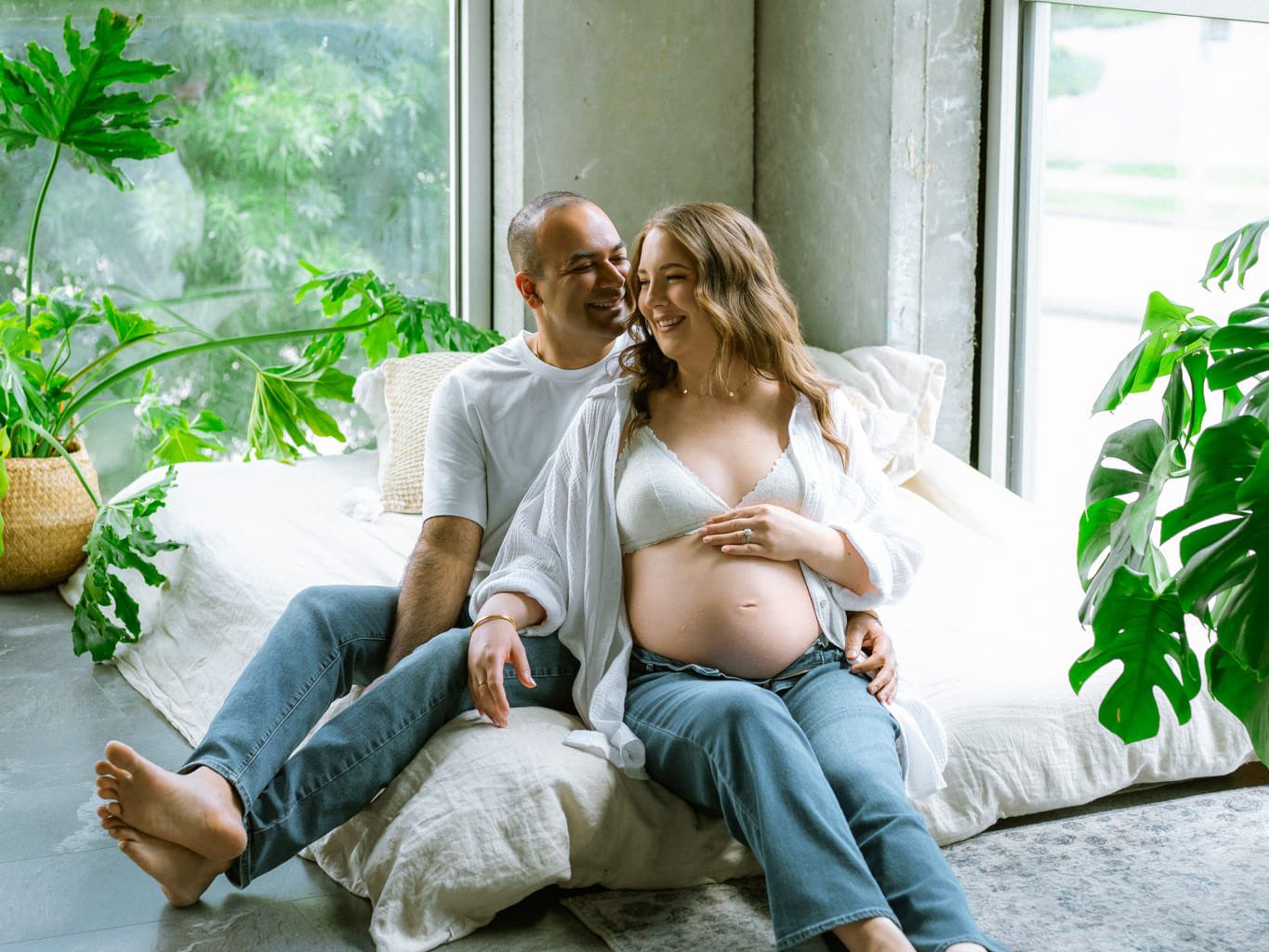 Maternity shoot studio session, expectant couple relaxing on cushions by a big studio window with plants