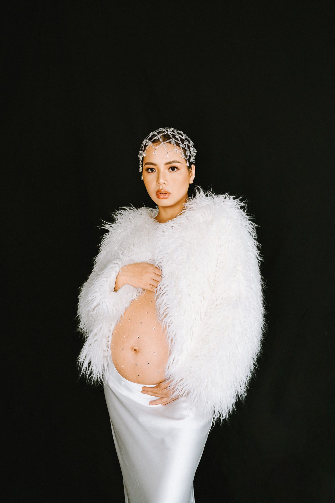 Maternity photoshoot in the studio, expectant mother in a white faux-fur jacket and crystal headpiece