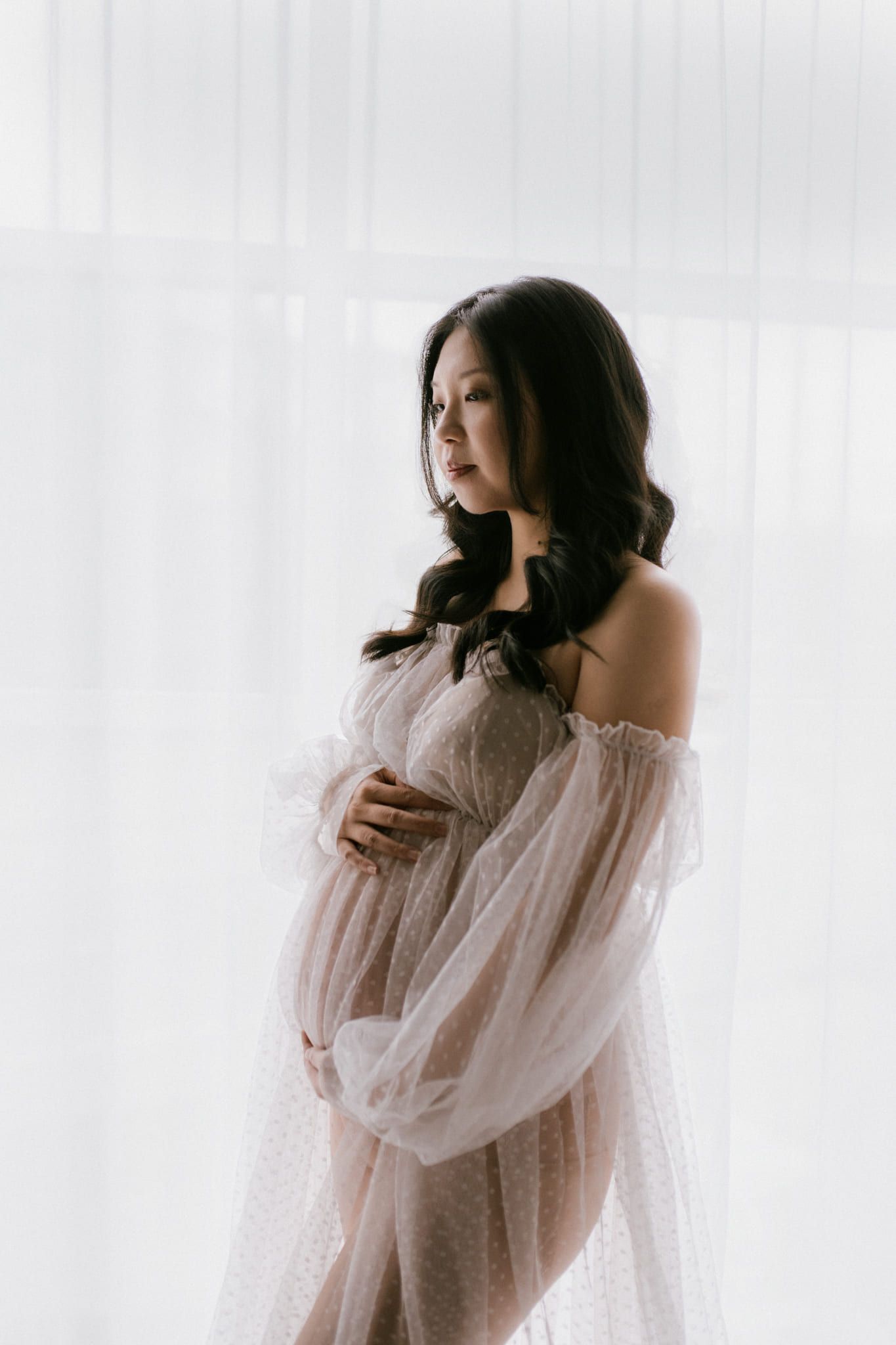 Maternity shoot in soft studio light, expectant mother in a sheer tulle gown