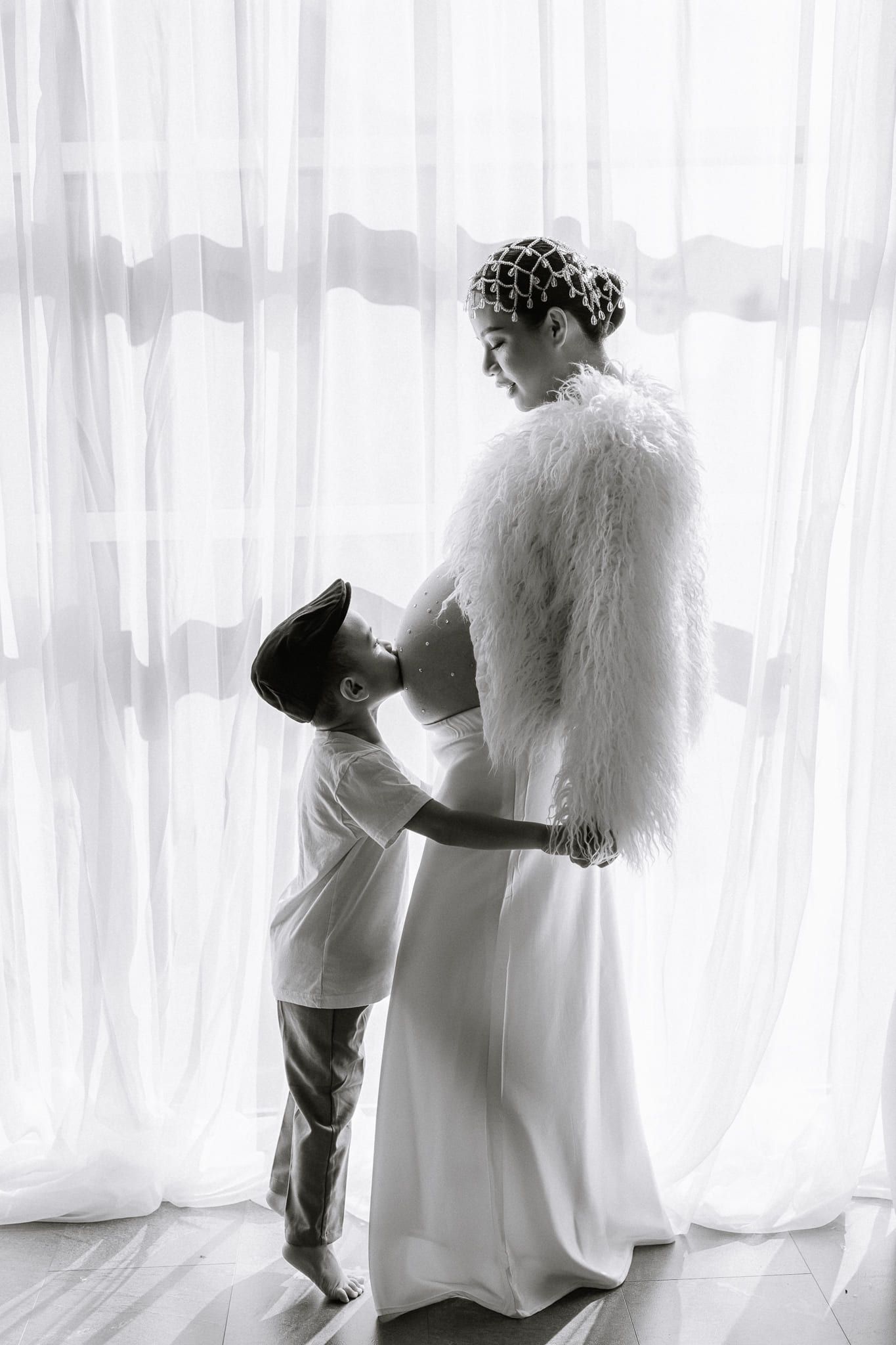 Expectant mother in a white faux-fur jacket and crystal headpiece in the studio, in black and white, by Coquitlam maternity photographer Natalie Varro