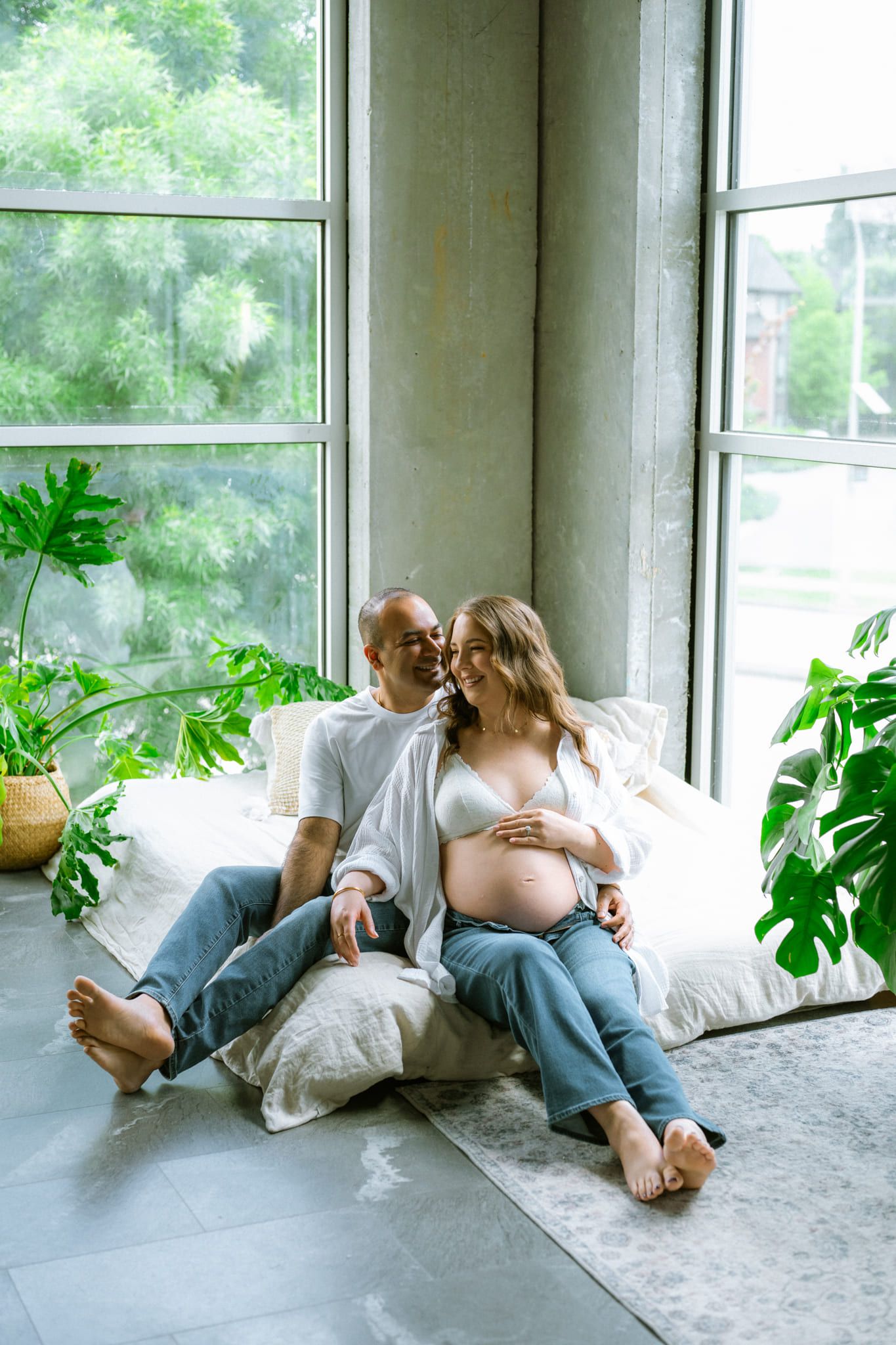 Indoor studio maternity photoshoot, expecting couple sitting together by the tall windows of a loft studio