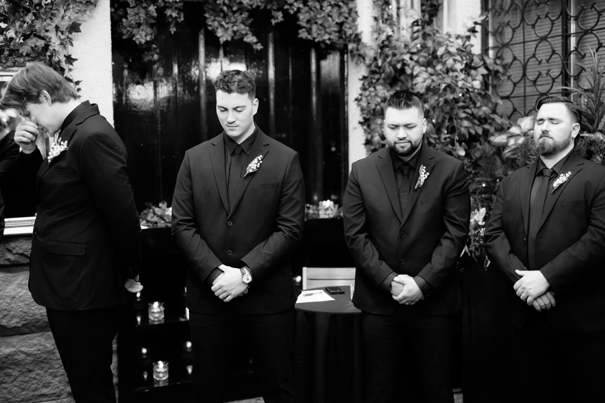 Black-and-white photo of the groomsmen in black suits before a dark doorway