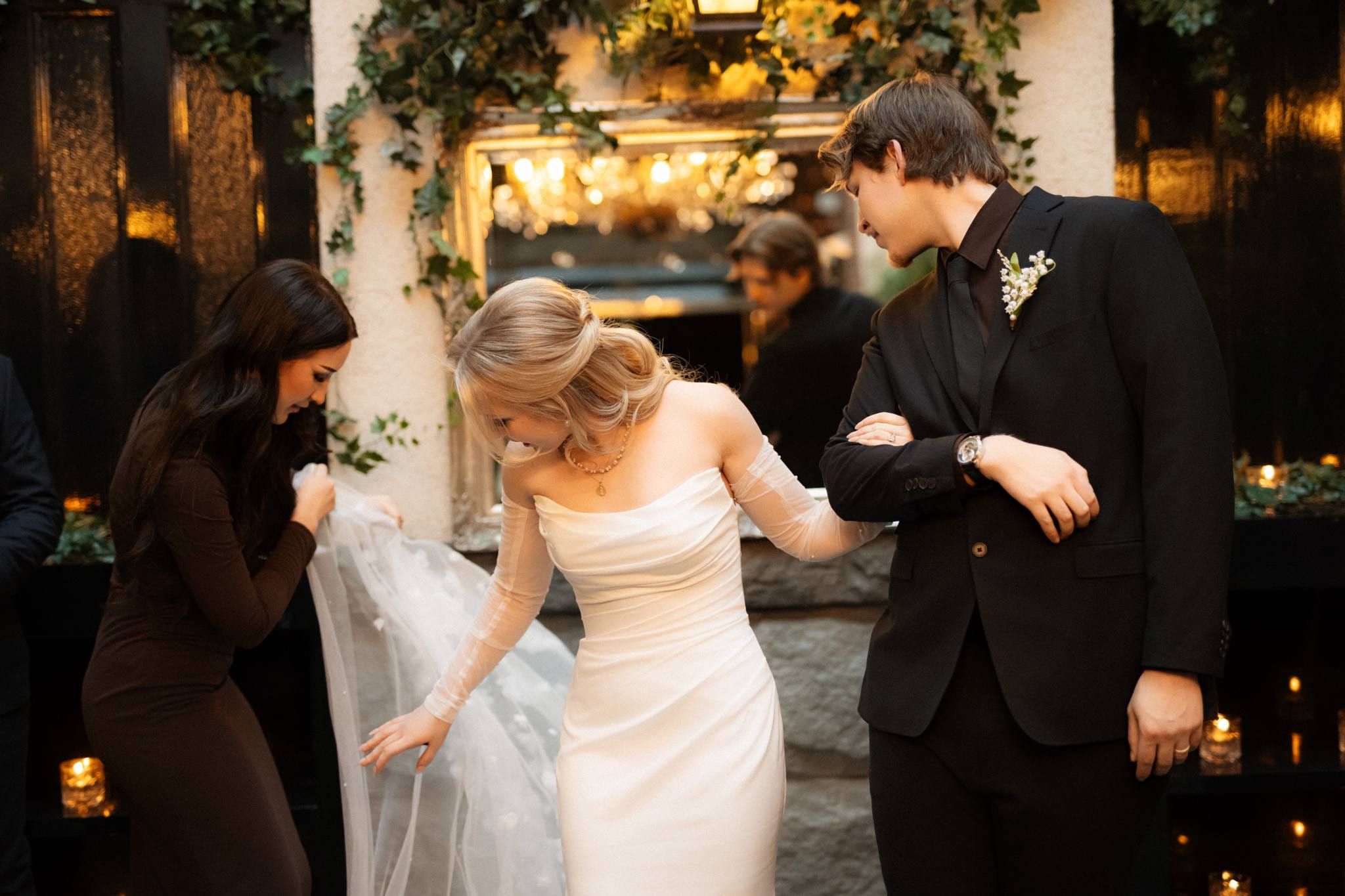 Bridesmaid adjusting the bride's veil while the groom holds her hand