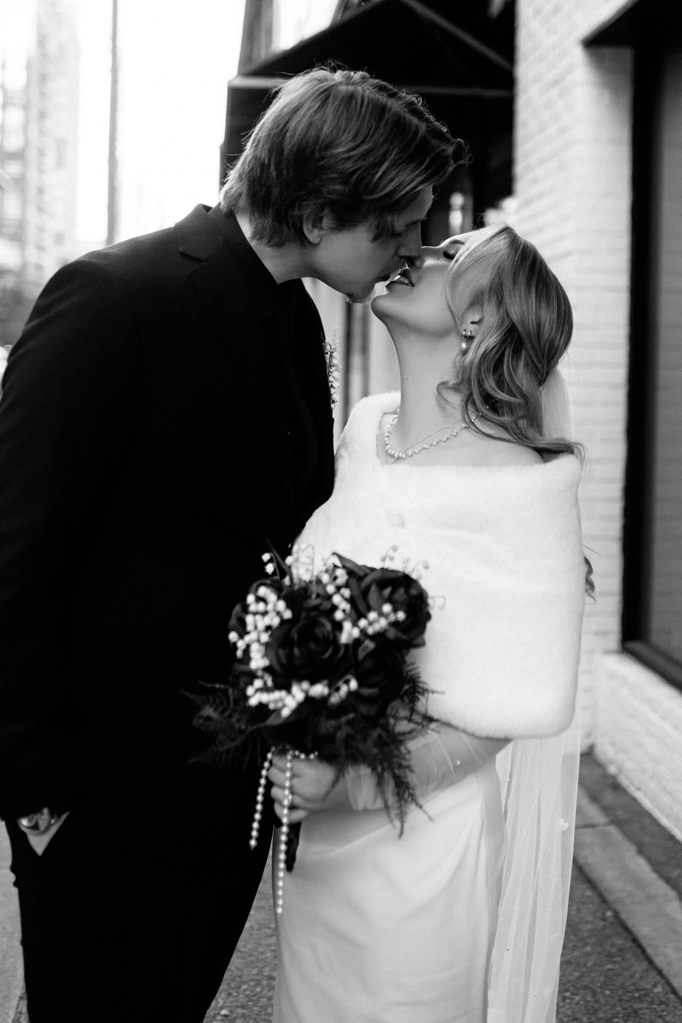 Black-and-white photo of the couple kissing on the street, bouquet in hand