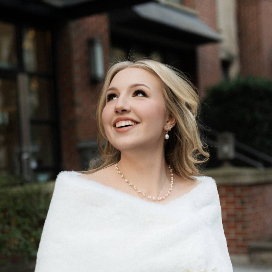 Viktoria in a white fur wrap smiling up before a brick building