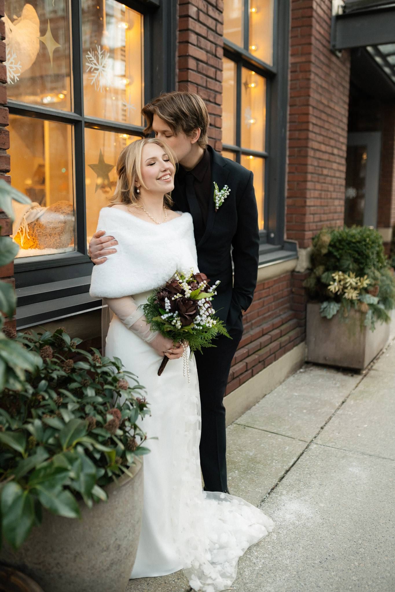 Bride in a white fur wrap with her bouquet beside the groom by a brick window