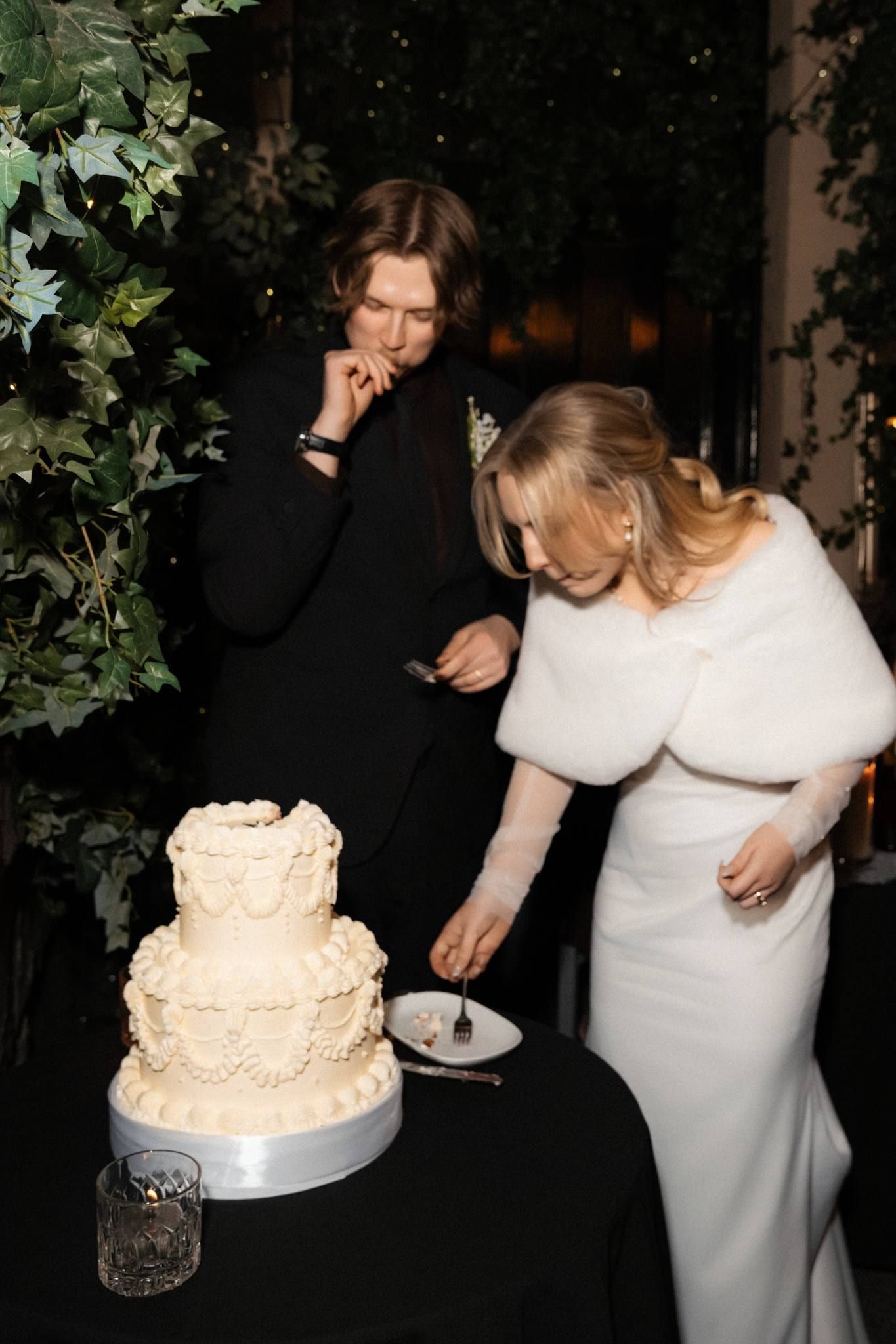 Groom looking on as the bride cuts the tiered wedding cake