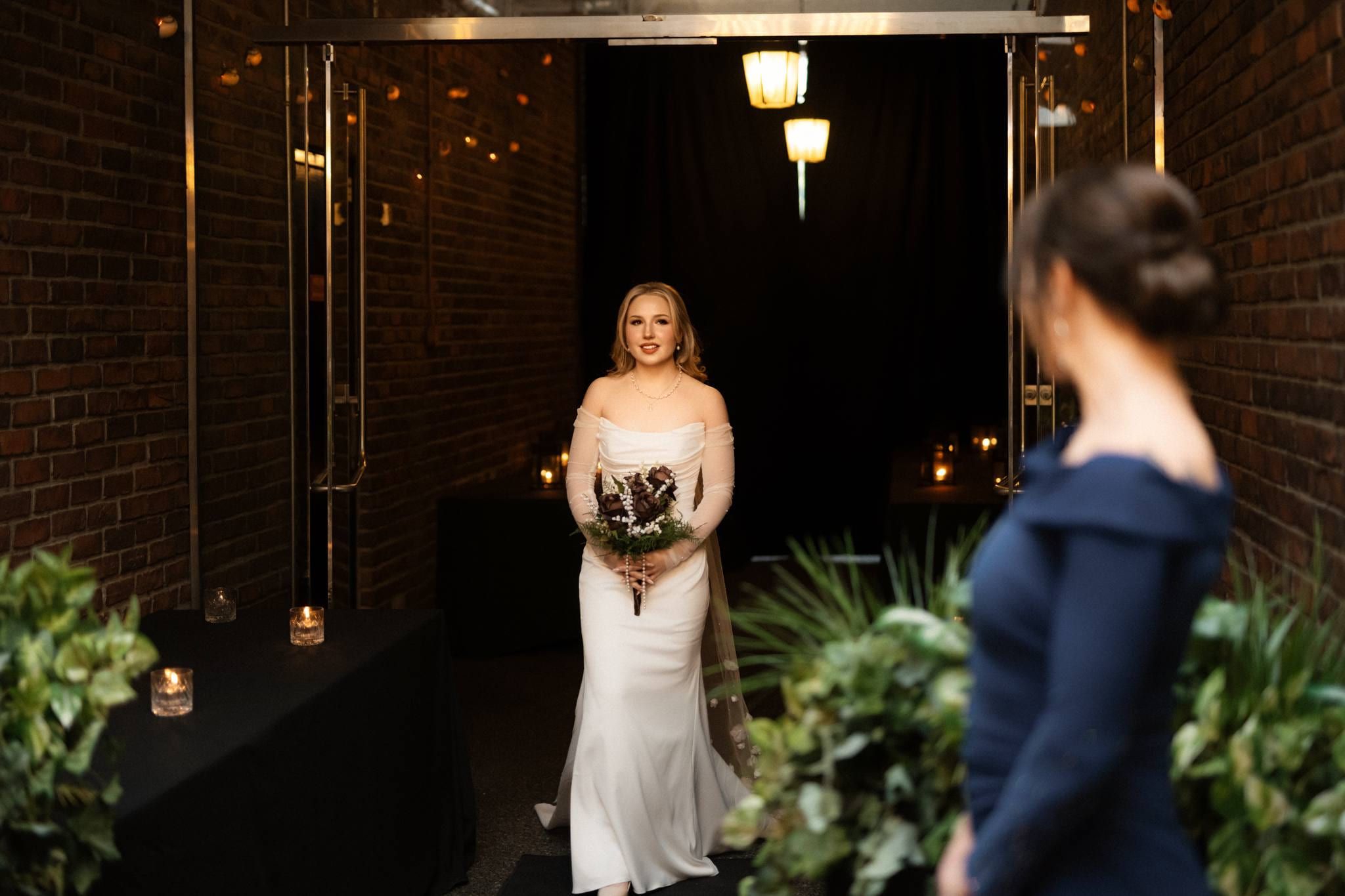 Bride holding her bouquet in a dark brick courtyard as a guest looks on
