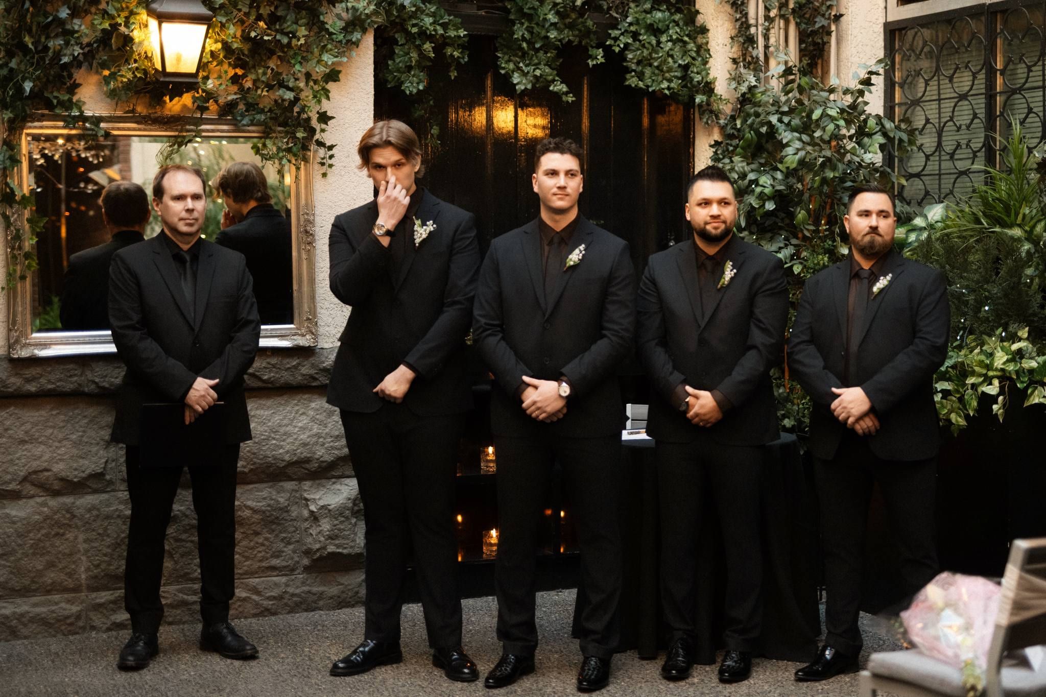 Groom and groomsmen in black suits lined up before an ivy-covered wall