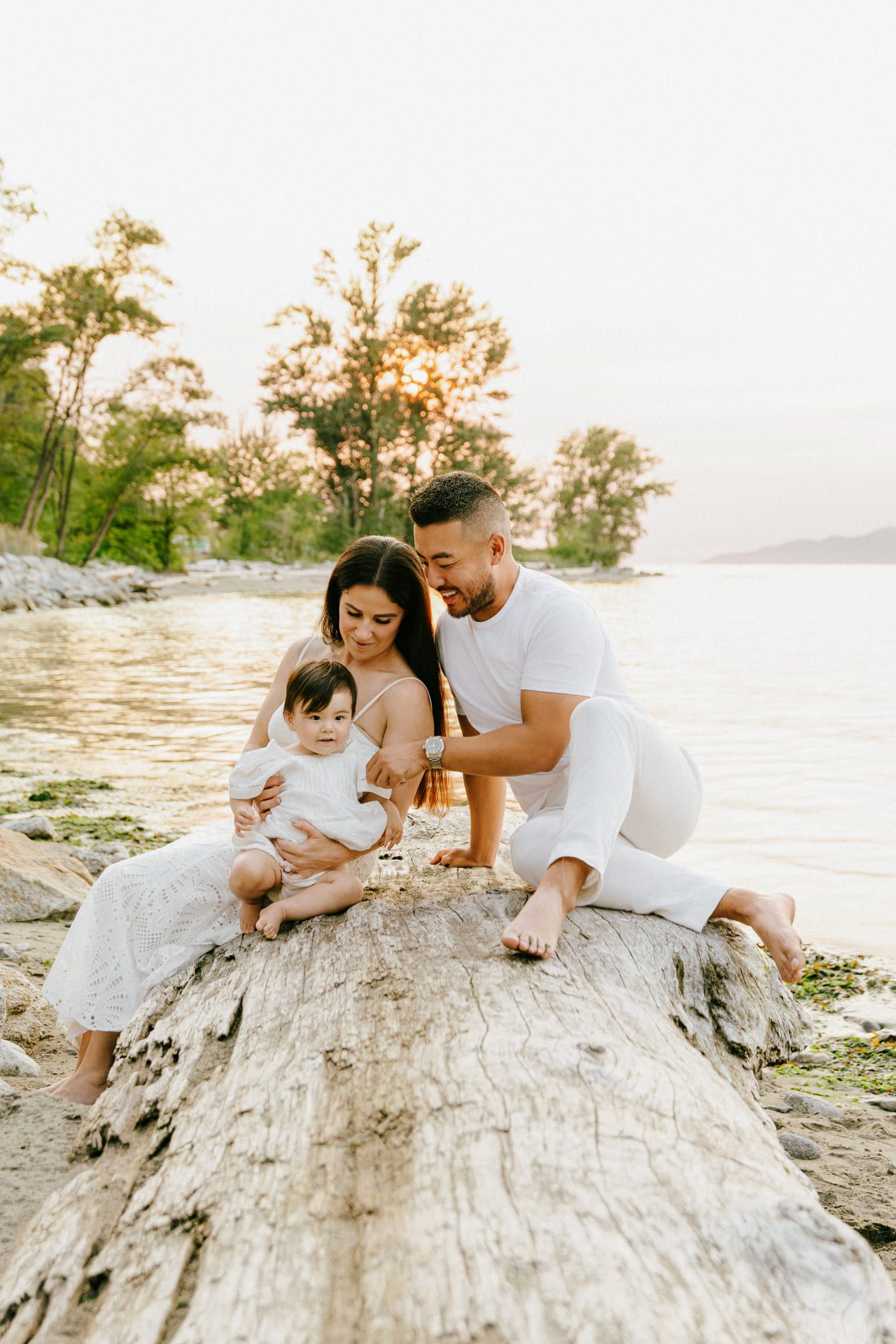 Professional family photography on a large driftwood log by the water, parents playing with their baby