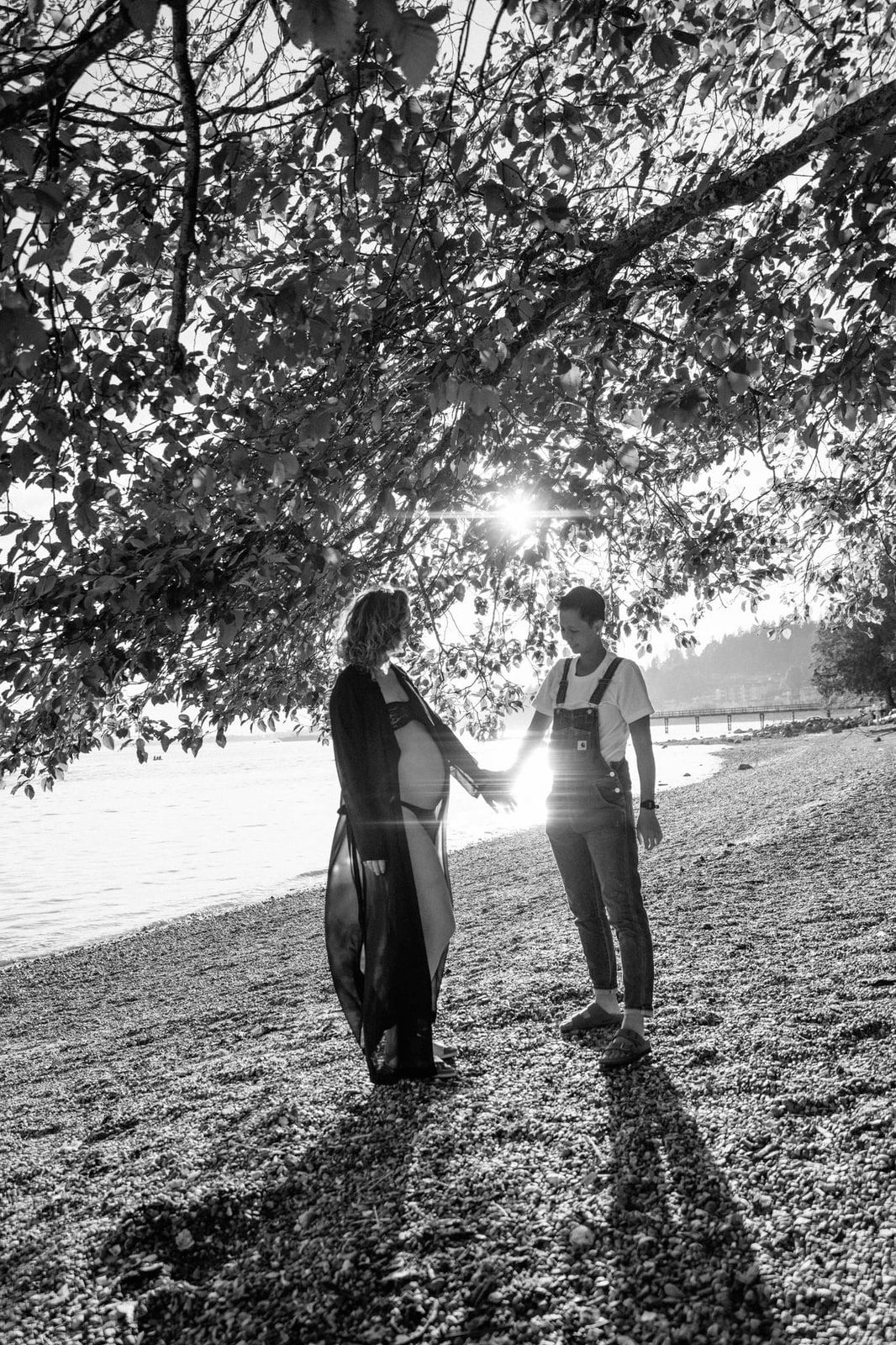 Black and white maternity shoot under a tree by the water, an expectant couple holding hands
