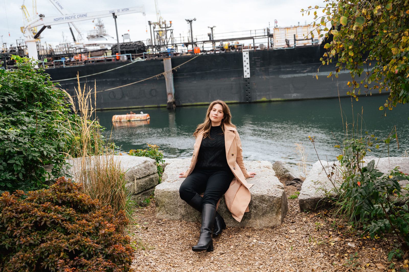 Portrait photoshoot, woman in a camel coat sitting on rocks by the waterfront