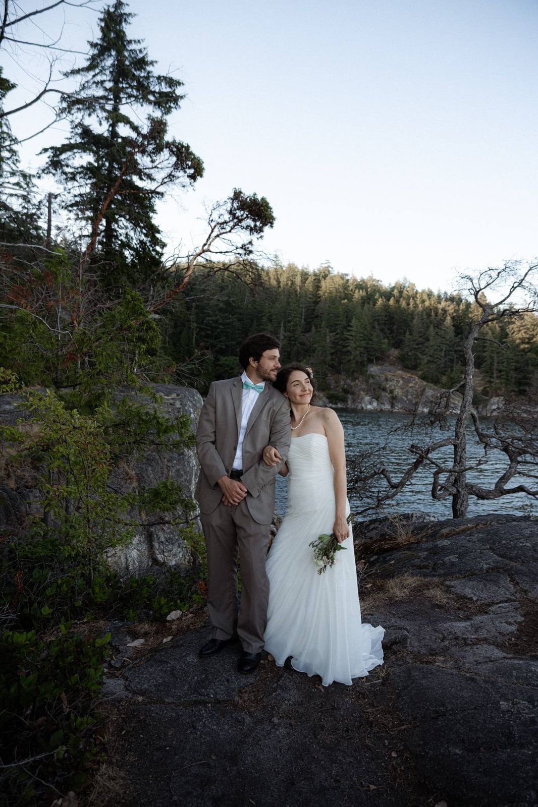 Elopement photoshoot, a couple in wedding attire stands close on a rocky bluff with evergreens and water behind