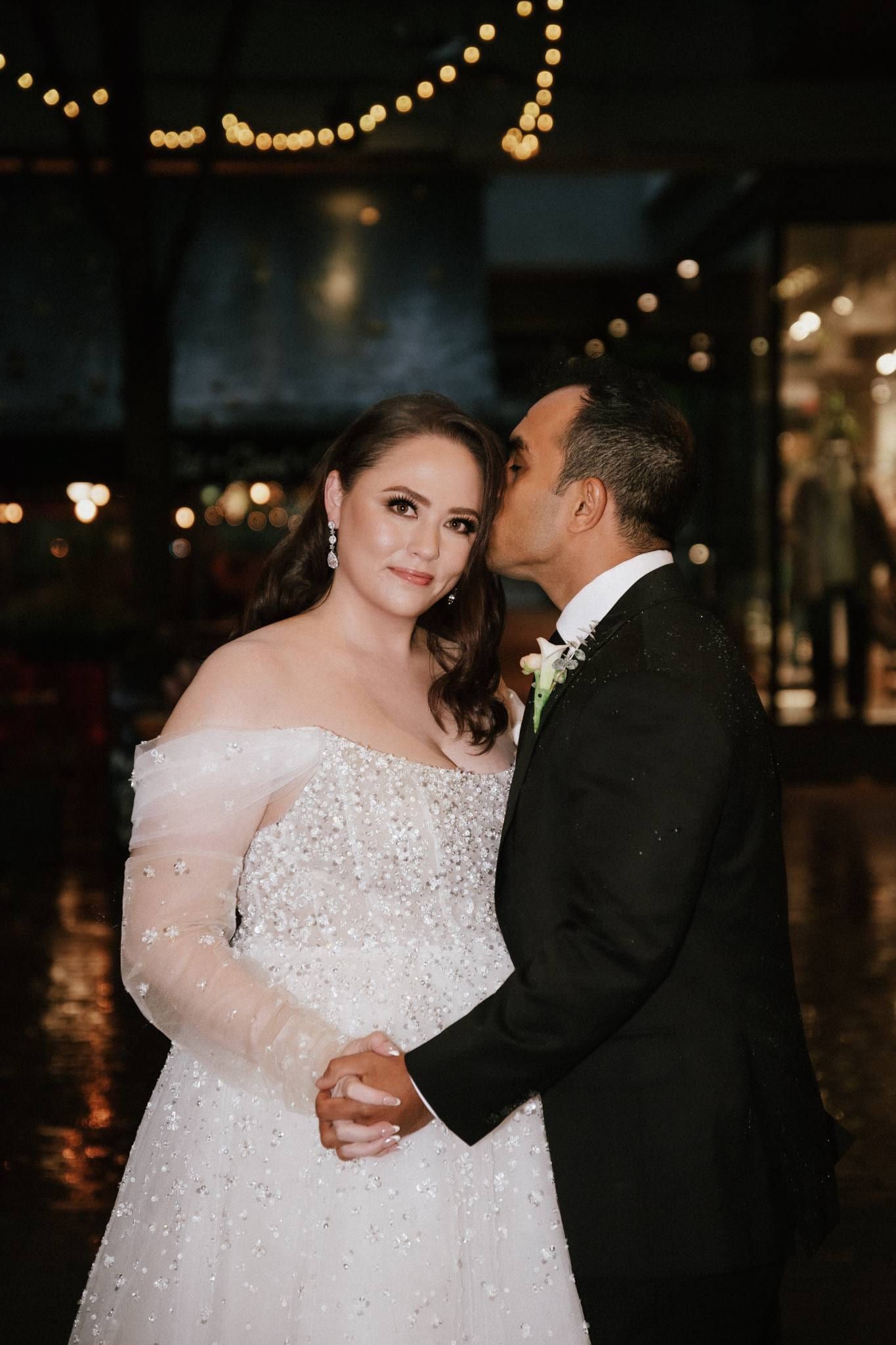 Professional wedding photos, groom kissing the bride's cheek under string lights in a dim venue