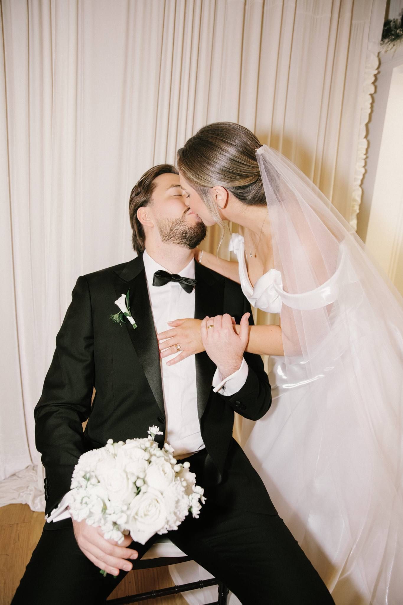 Indoor wedding photography, groom in a black tuxedo kissing the bride's forehead, bouquet in hand, by sheer curtains