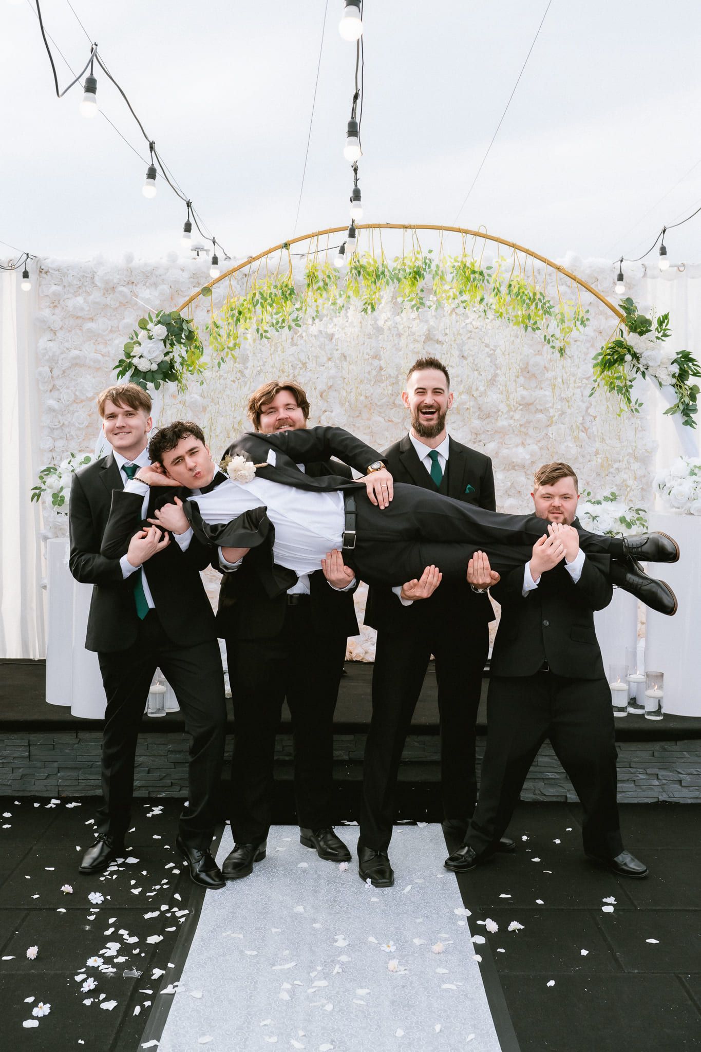 Groom photography in front of a floral arch, groomsmen lifting the groom