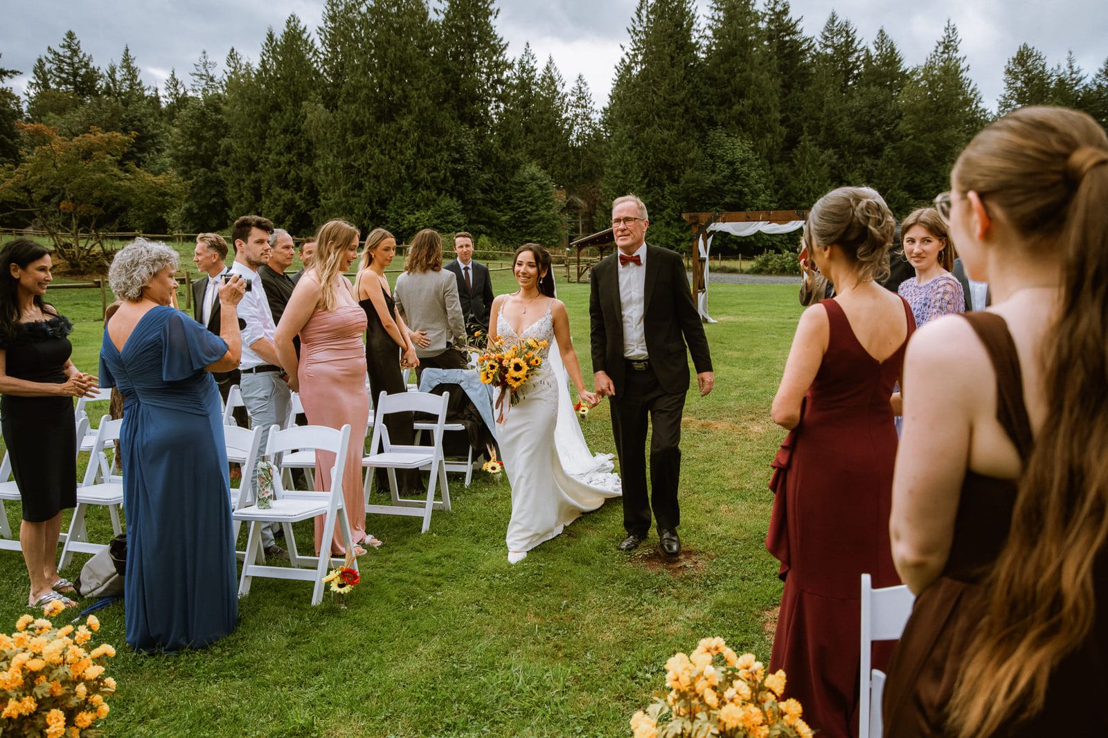 Wedding photography in Port Moody, bride walking down a grassy aisle as guests stand and turn
