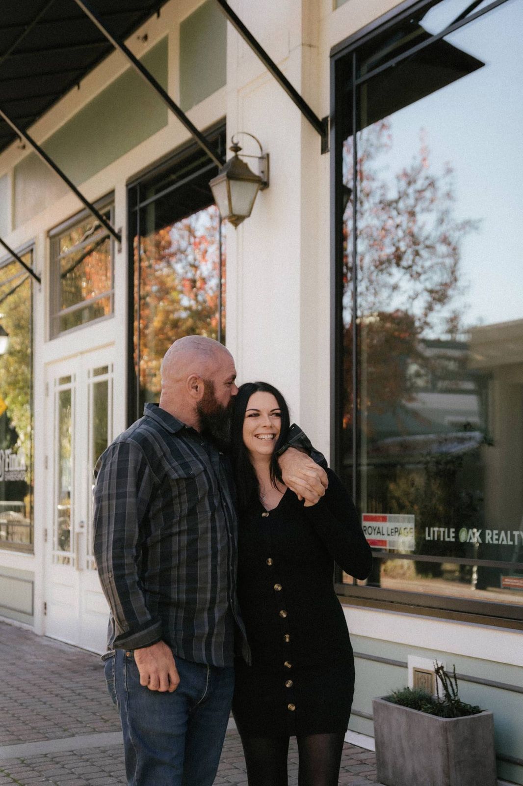 Indoor engagement pictures, a couple smiles in an embrace outside a storefront with tall windows and lanterns