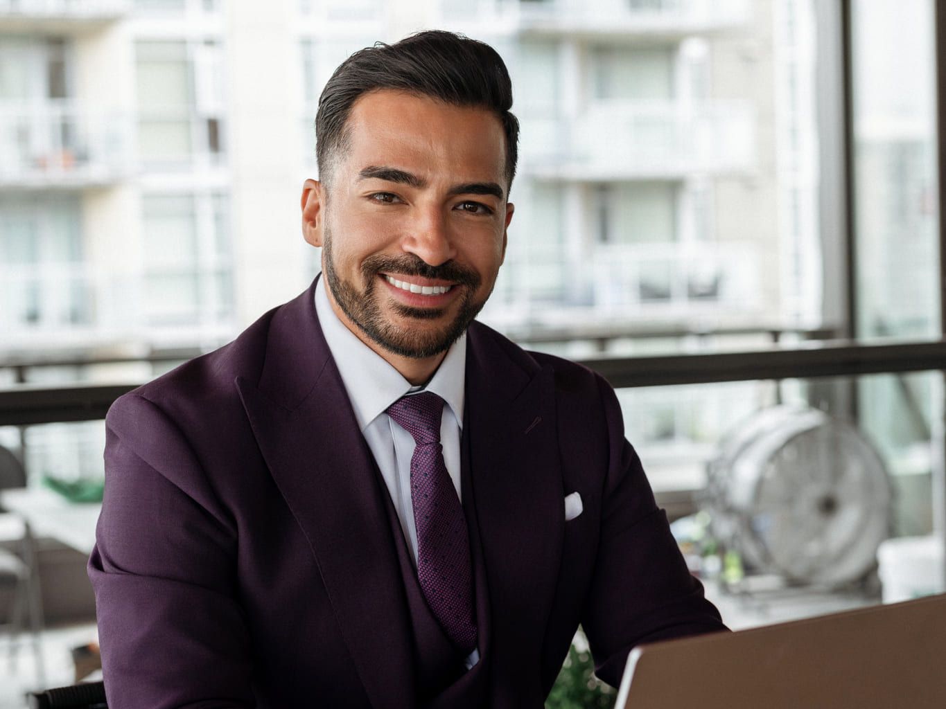 Coquitlam headshot photographer, smiling man in a plum suit and tie seated by an office window