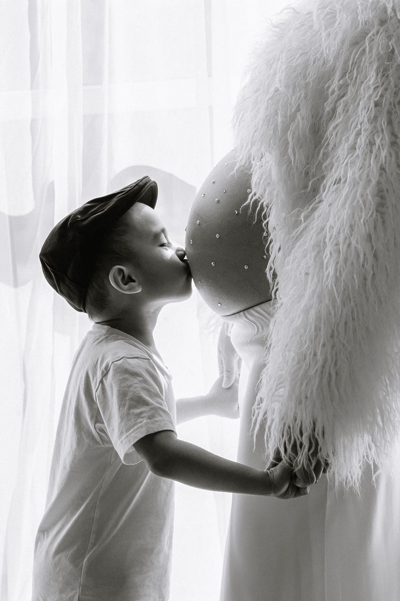 Family maternity photography by a window, little boy kissing his mom's baby bump, in black and white