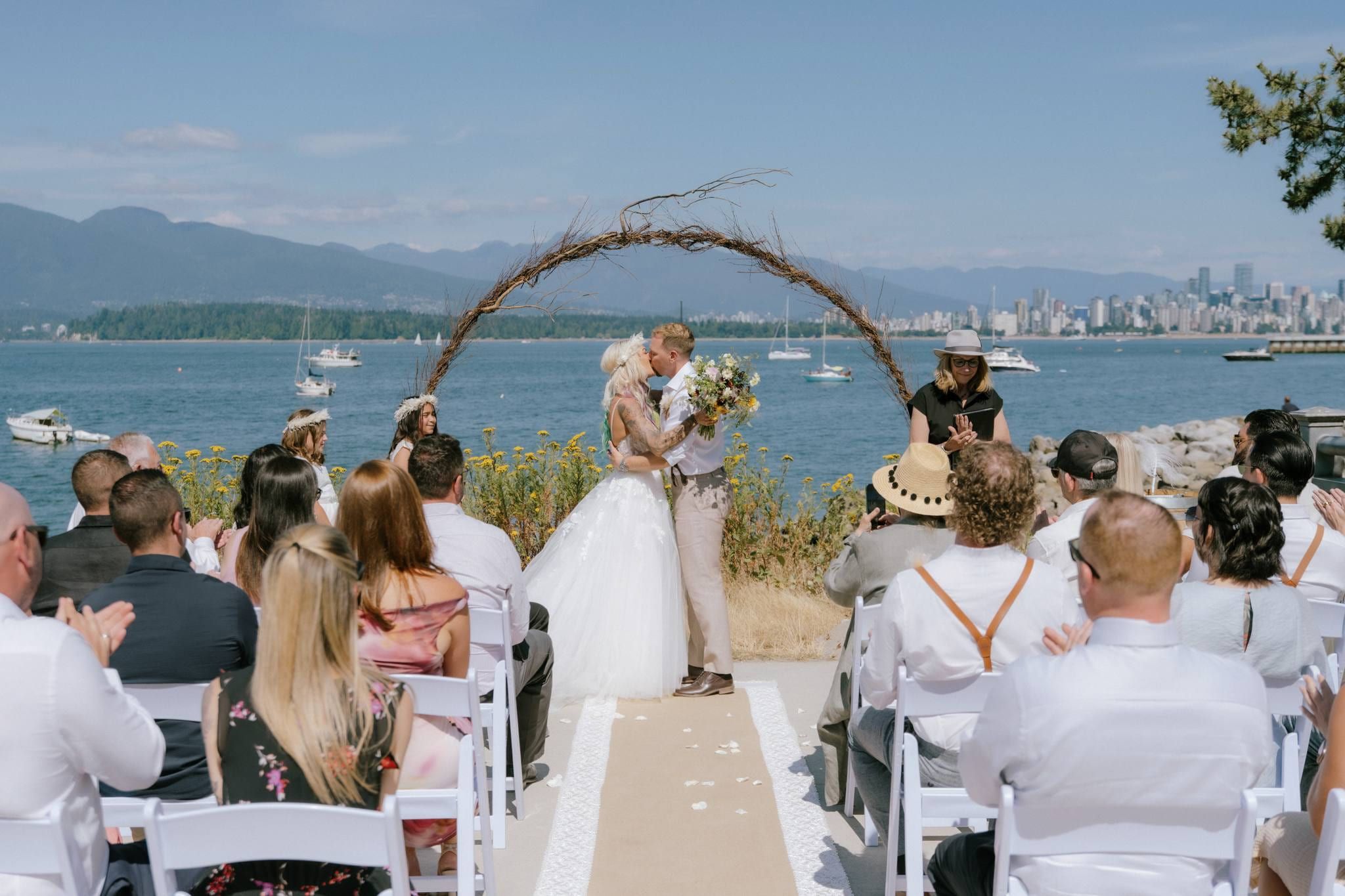 Whitney and James kissing beneath a driftwood arch with the water and city skyline behind