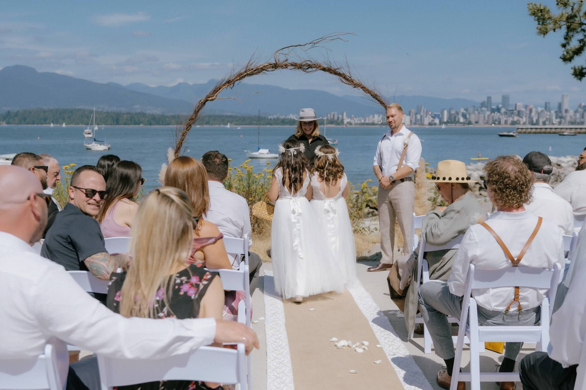 Guests watching the couple beneath a driftwood arch with the city skyline behind
