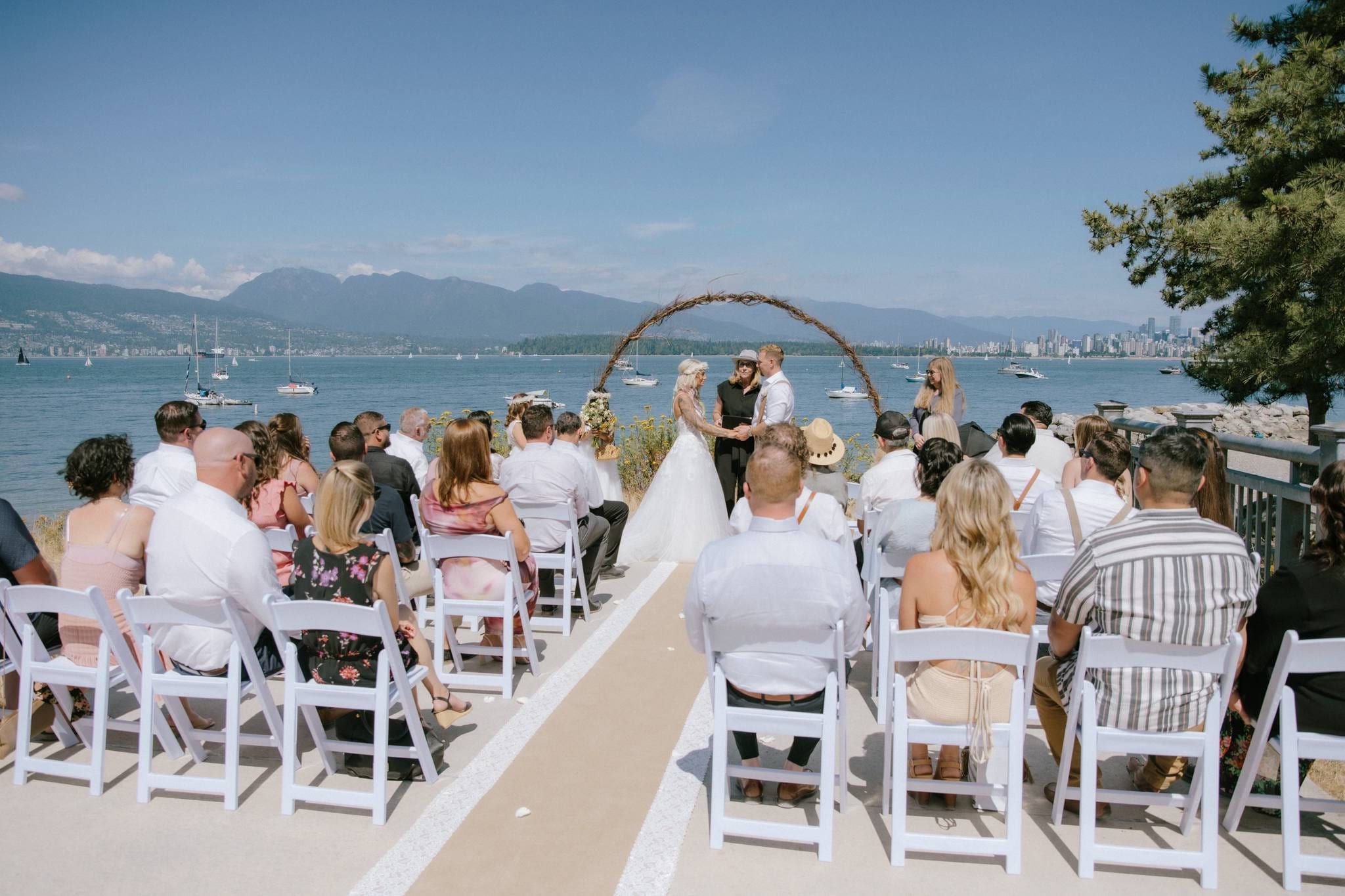Guests seated before a driftwood arch at the beach ceremony with the city skyline behind