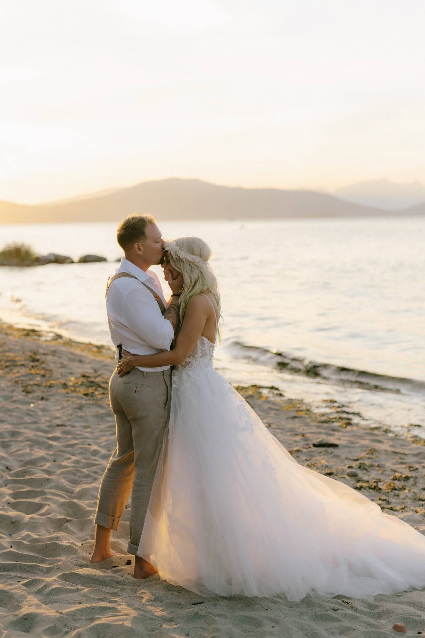 Groom kissing the bride's forehead on the beach at sunset