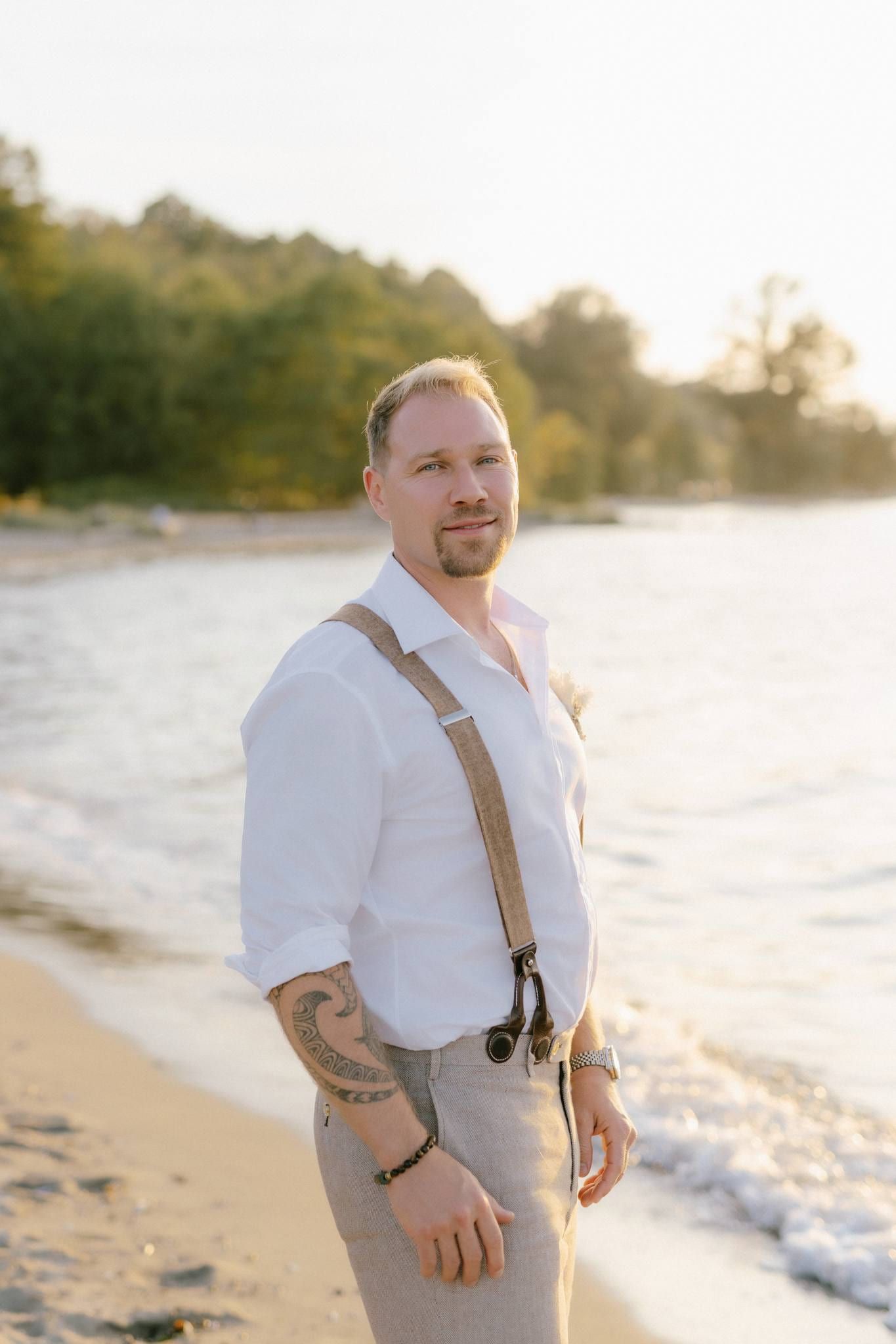 Groom in a white shirt and suspenders standing on the beach