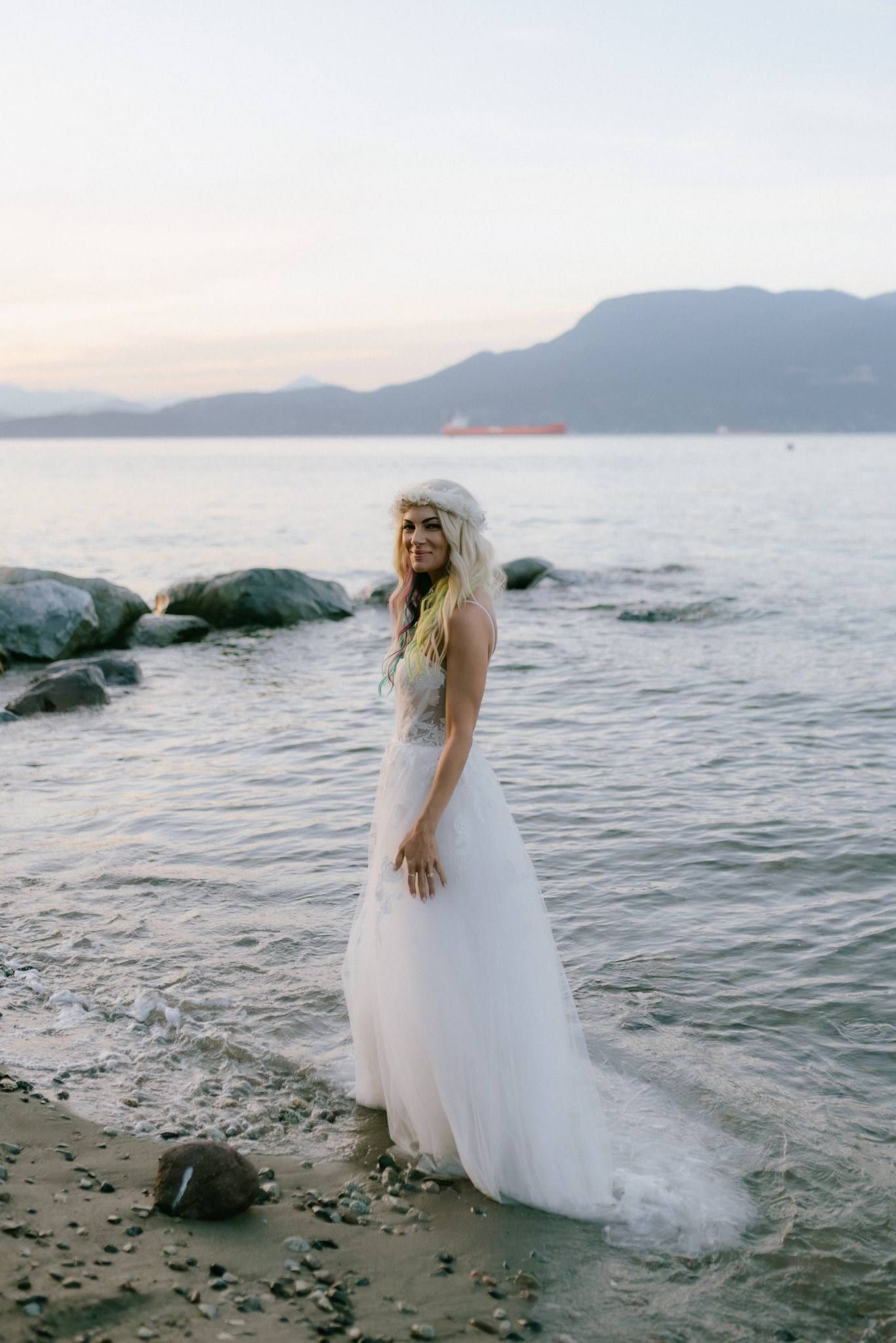 Bride standing on rocks at the water's edge with mountains beyond
