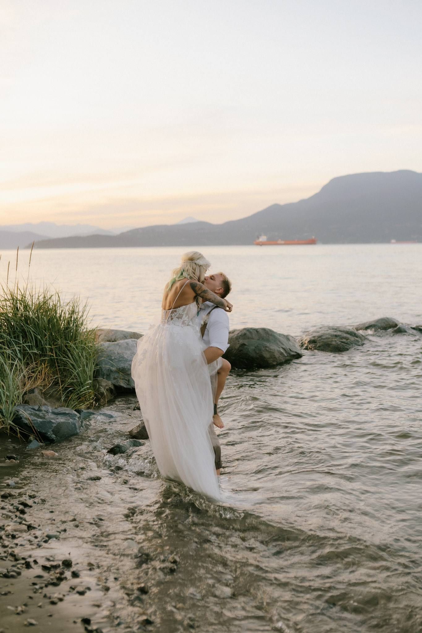 Bride standing on rocks at the water's edge, mountains beyond