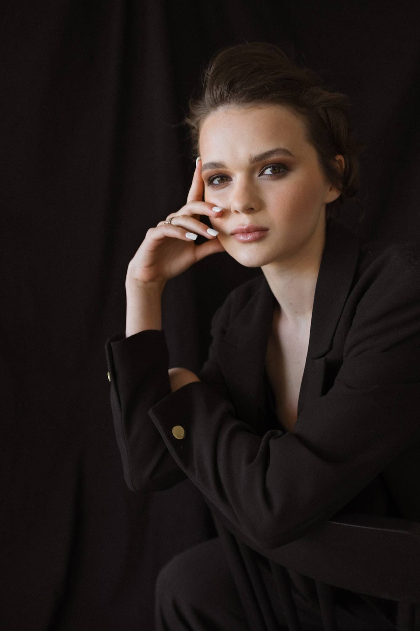 Model in a black blazer seated on a studio block, hand at her chin